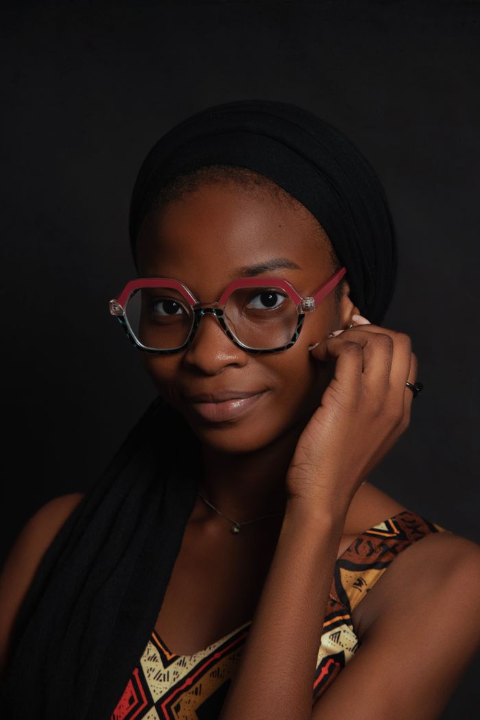 Studio portrait of a stylish woman in a hijab and glasses against a black background.